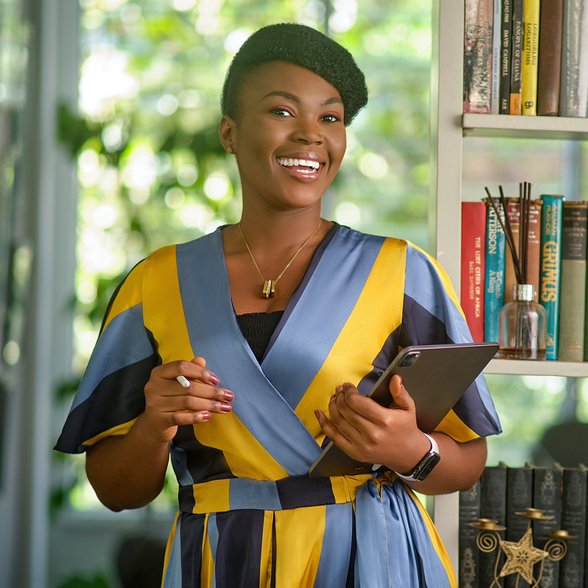 A person stands indoors holding a tablet and a pen. They are wearing a dress with blue, yellow, and black stripes. A bookshelf with books and decorative items is visible in the background, and natural light streams in through large windows.