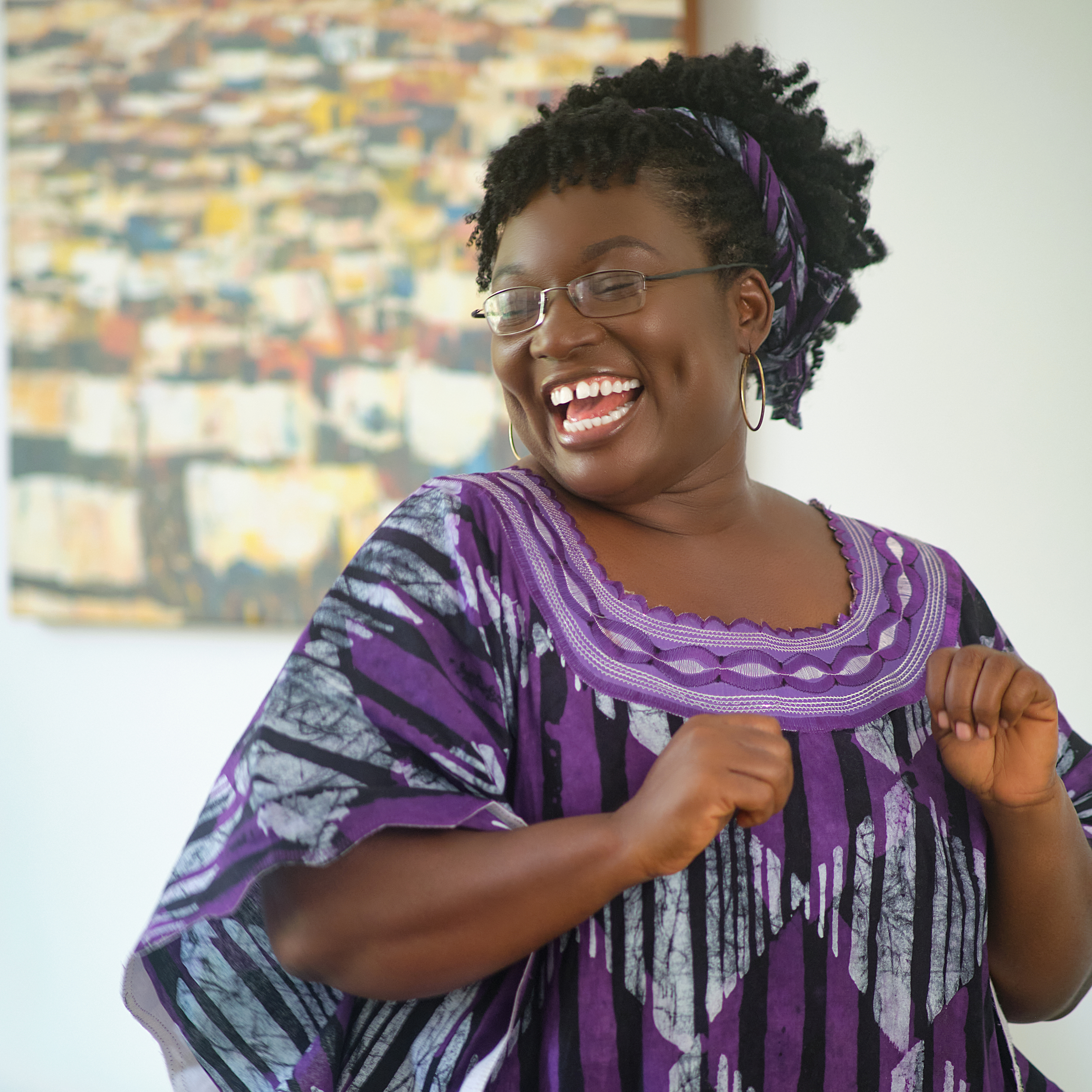 A person wearing a purple and black patterned traditional outfit with a matching headscarf stands in front of an abstract painting.